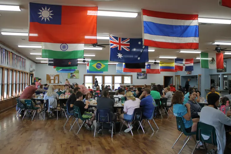 The image depicts many people seated at round tables enjoying a meal. Along the ceiling hangs flags representing different countries around the world.