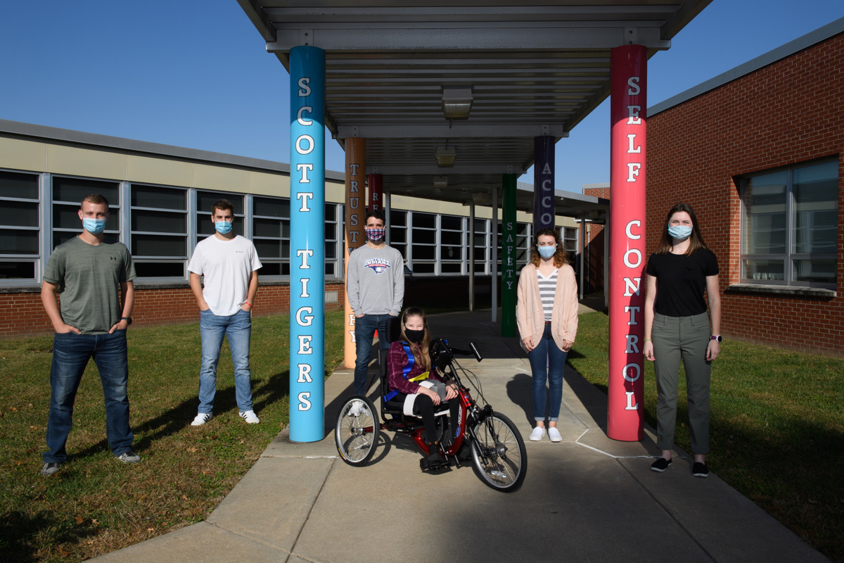 Charleigh Garrett poses with the five USI students (now alumni) who designed and developed the adaptive tricycle she is on in front of Scott Elementary School