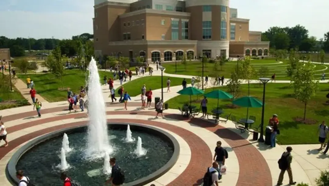 Various students walk alongside the fountain from a top-down view, with Rice Library in the distance.