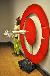 Sculpture of a person with an apple on their head, a large target, and a dart in the bullseye