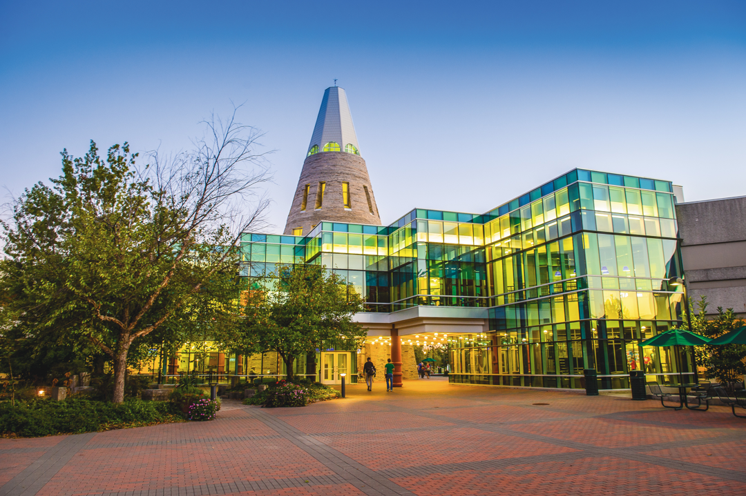 A view of the University Center, including the Tower, at dusk with the lights on green trees