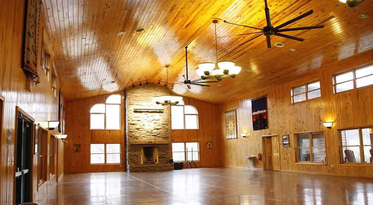 The image depicts a large meeting space with wood floors and walls and large windows. The room is empty.