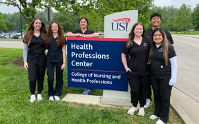 Graduating Respiratory Therapy students pose together in scrubs by the Kinney College of Nursing and Health Professions sign